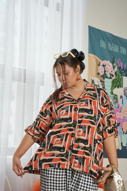 Model standing and admiring the breezy feel of Buttoned Belle Blouse in Abstract Garden, lifting shirt with a relaxed expression, featuring abstract print and loose fit.